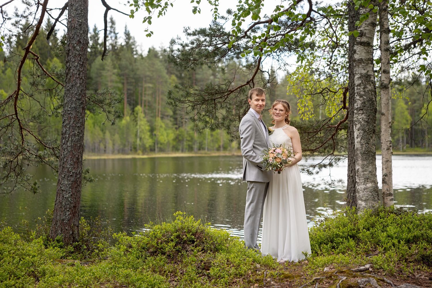 Hääpari seisoo lammen rannalla metsämaisemassa.