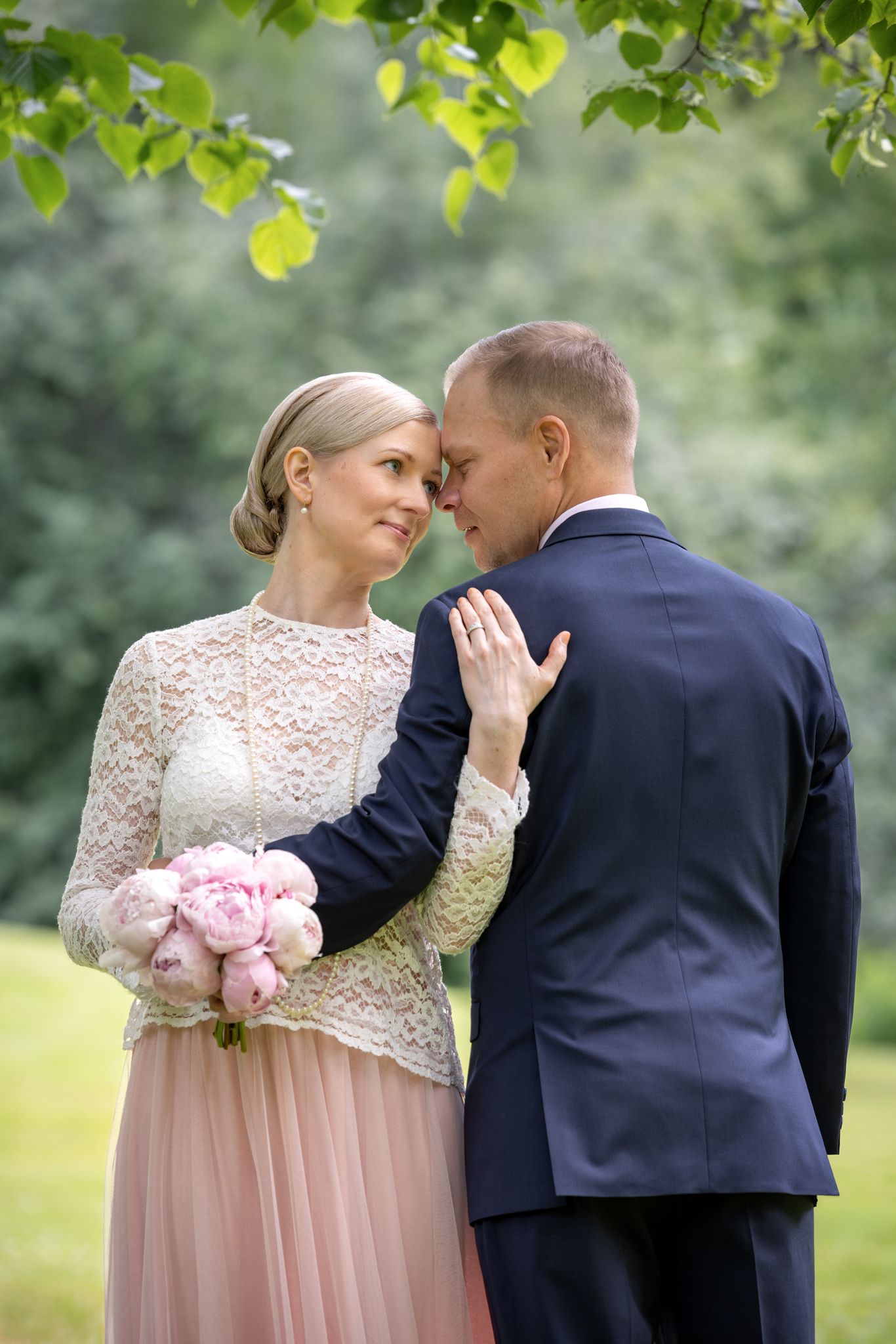 Bride in lace dress holds pink peonies while touching groom's face in navy suit, sharing intimate moment outdoors.