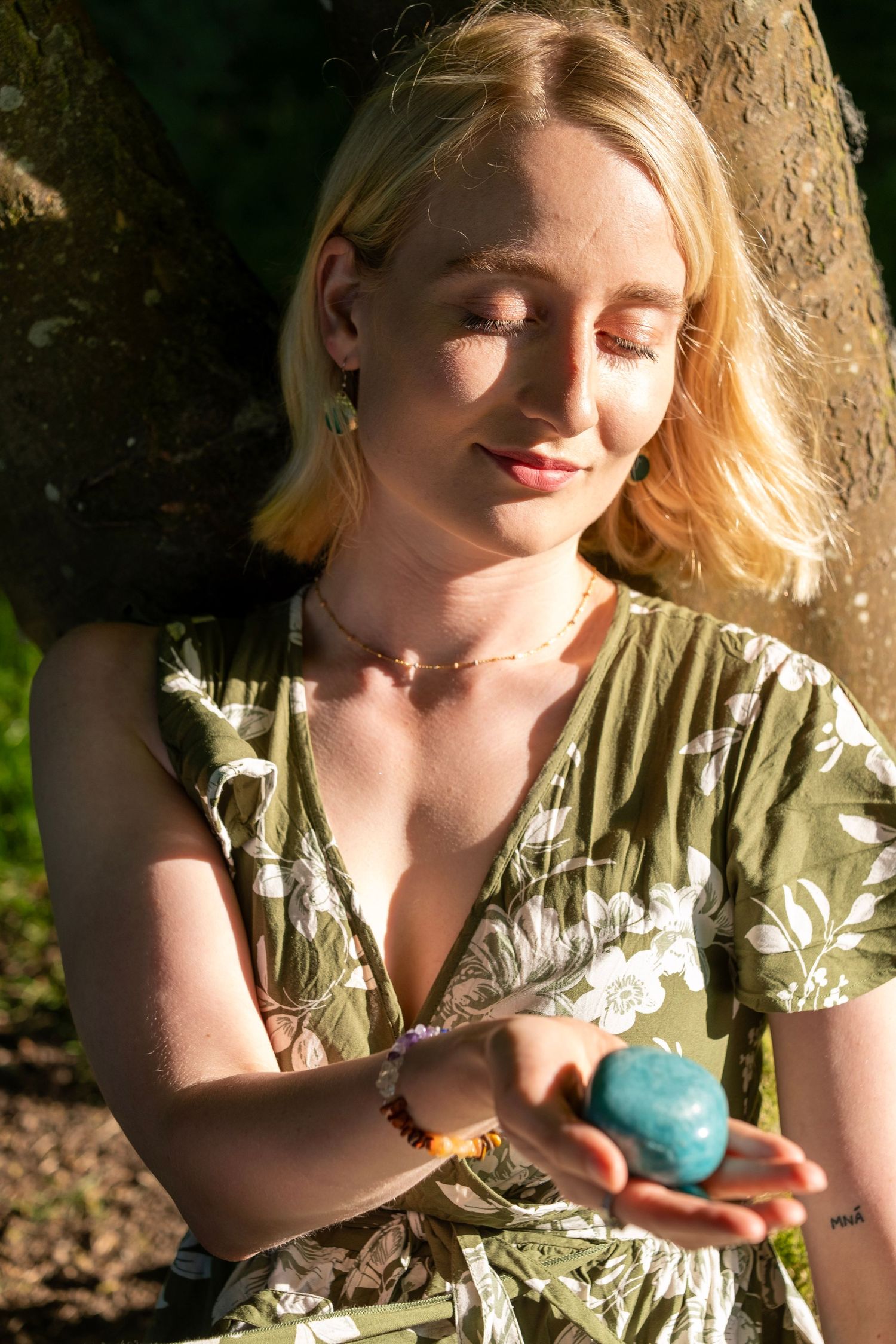 A person in a floral dress holds a blue crystal sphere while sitting in dappled sunlight against a tree trunk.