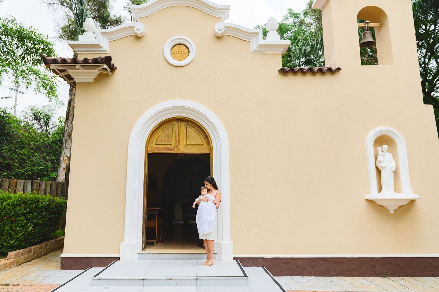A small tan Spanish colonial style chapel with white trim features an arched entrance doorway and decorative niche.