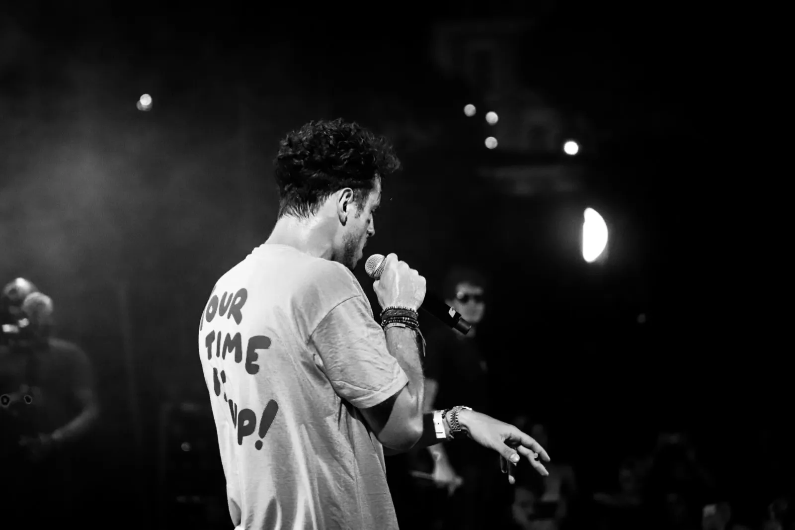 Rappeur en t-shirt clair, capturé en contre-plongée, micro à la main sur une scène éclairée par des faisceaux lumineux dans la pénombre.
