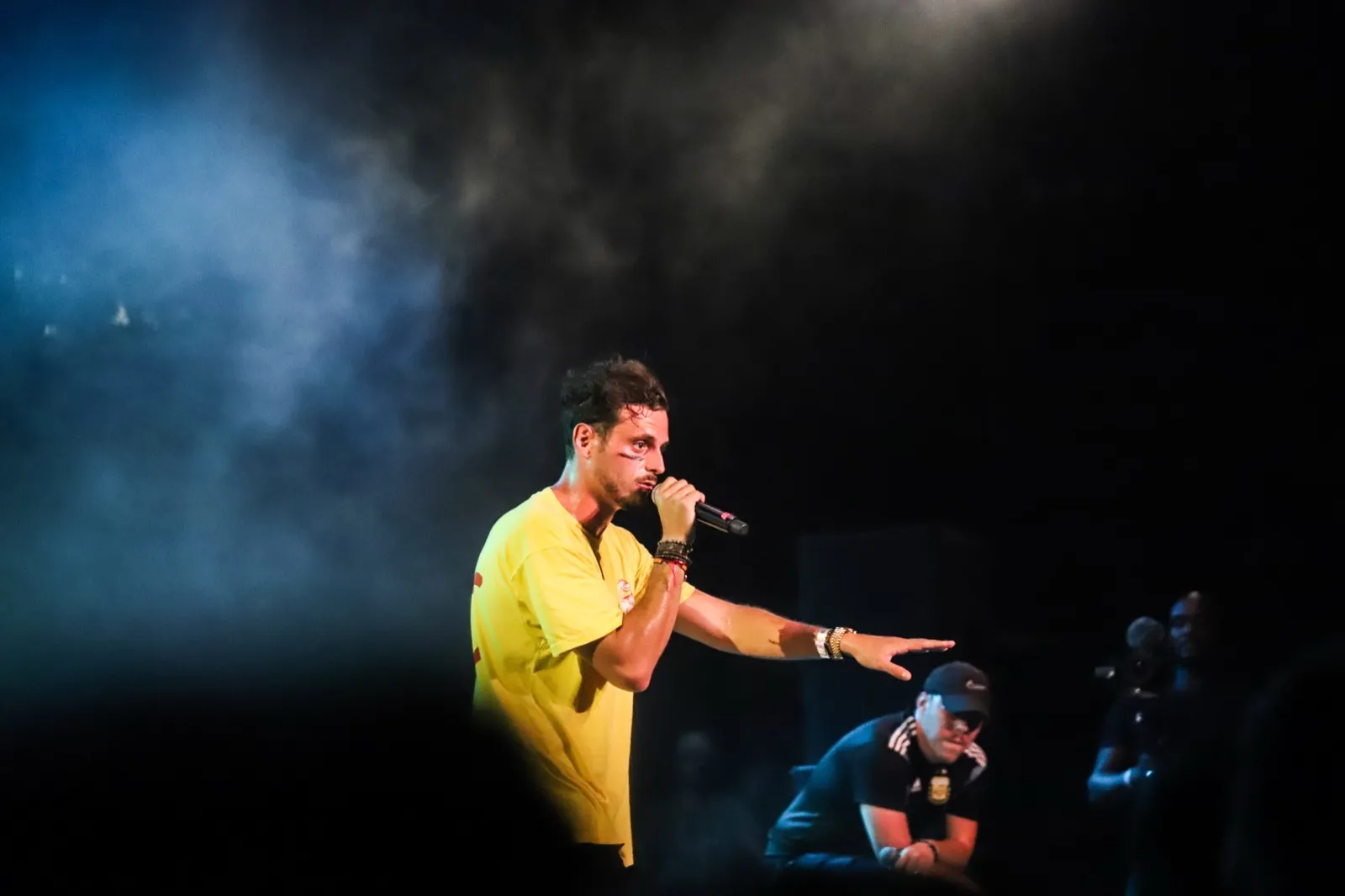 Chanteur habillé en jaune fluo, micro en main, penché vers le public avec intensité, entouré de brume et d’éclairage dramatique.