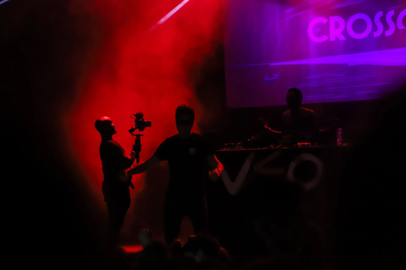 Scène de concert sous lumière rouge, un vidéaste capture la performance d’un artiste, pendant qu’un DJ joue à l’arrière-plan.