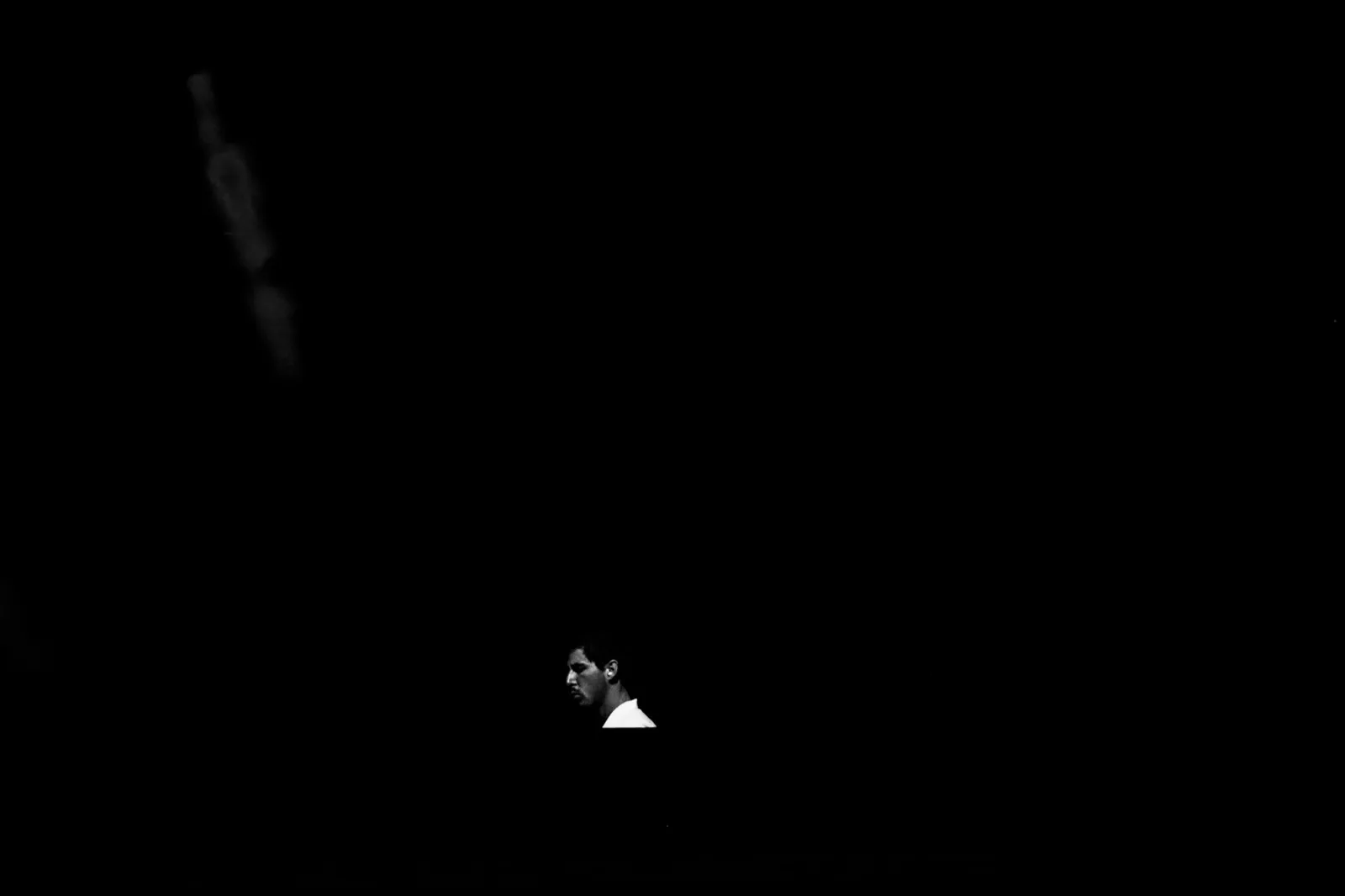 Artiste isolé sur une scène plongée dans l’obscurité totale, seul un spot blanc éclaire sa silhouette minuscule.