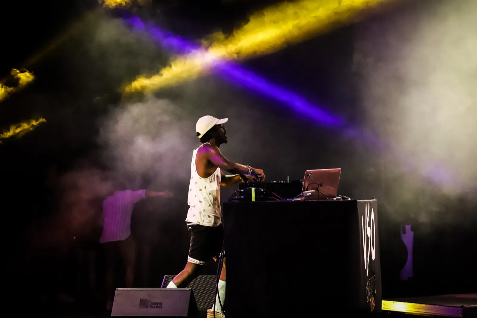 MC sur scène à côté de sa table de mixage, entouré de fumée et de faisceaux lumineux violets et jaunes, ambiance électrisante.