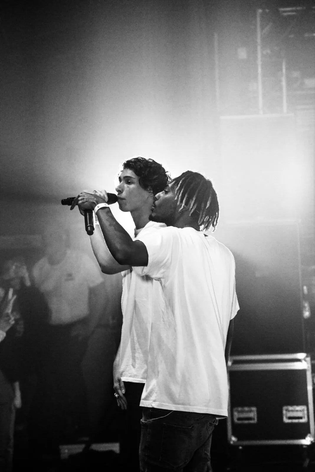 Un rappeur en t-shirt blanc sur scène, micro à la main, chantant face à une foule dans une lumière tamisée, avec un second rappeur.