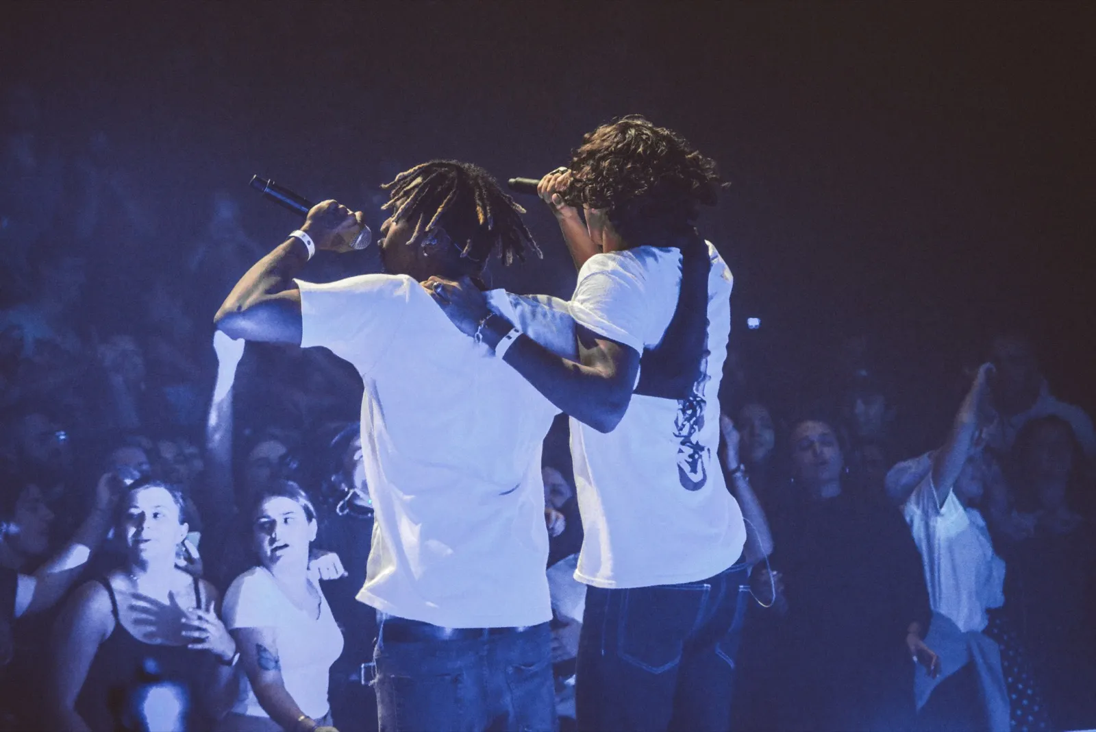 Moment de complicité entre deux chanteurs en plein concert, sous des lumières bleutées, entourés de fans enthousiastes.