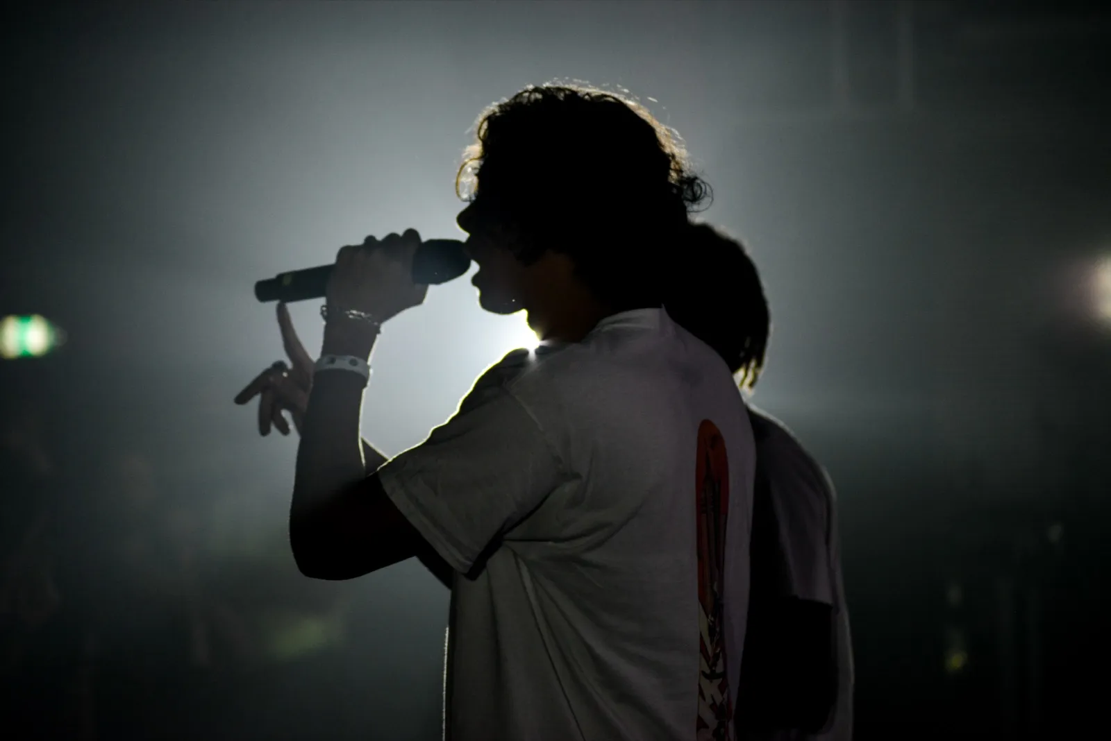 Silhouette d’un chanteur en t-shirt blanc, micro près de la bouche, baignée dans un éclairage tamisé et dramatique.