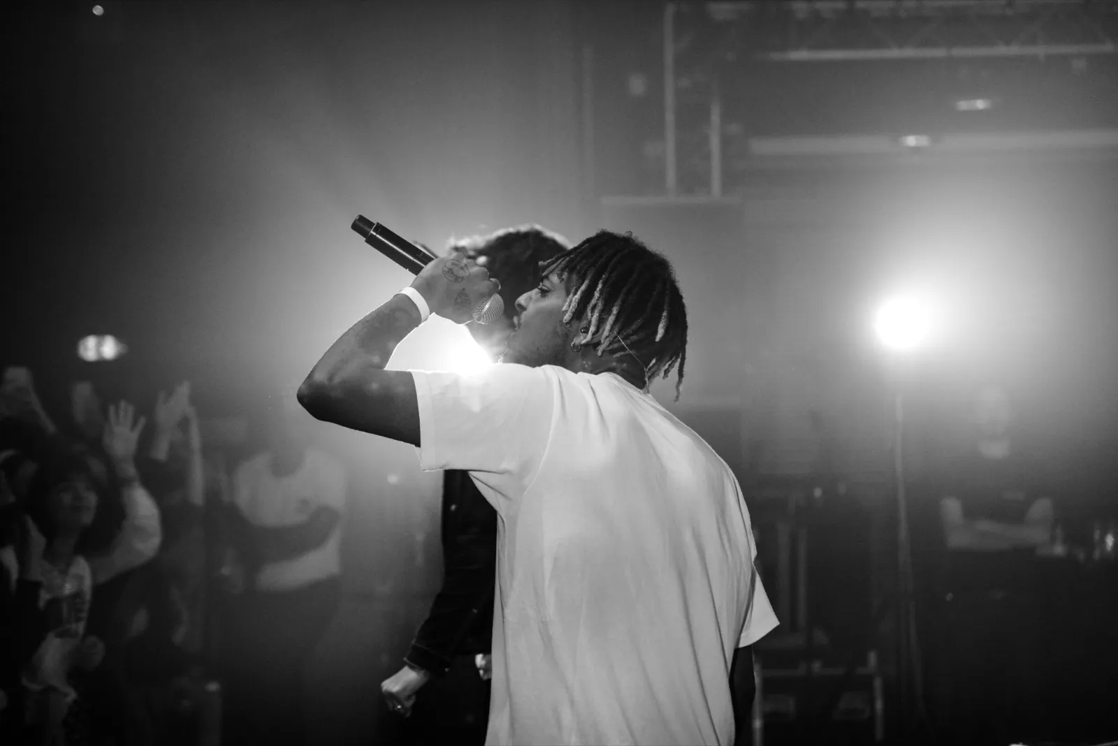Rappeur sur scène en noir et blanc, vu de dos, tenant un micro devant une foule dans une atmosphère brumeuse et contrastée.