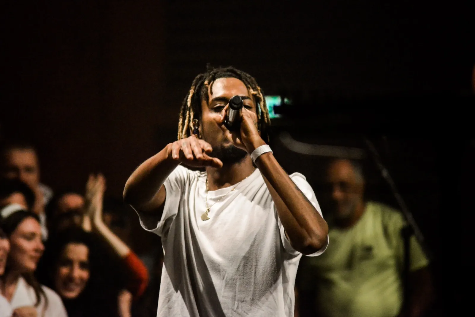 Artiste sur scène en t-shirt blanc, pointant du doigt le public avec intensité, éclairé par une lumière de concert tamisée.