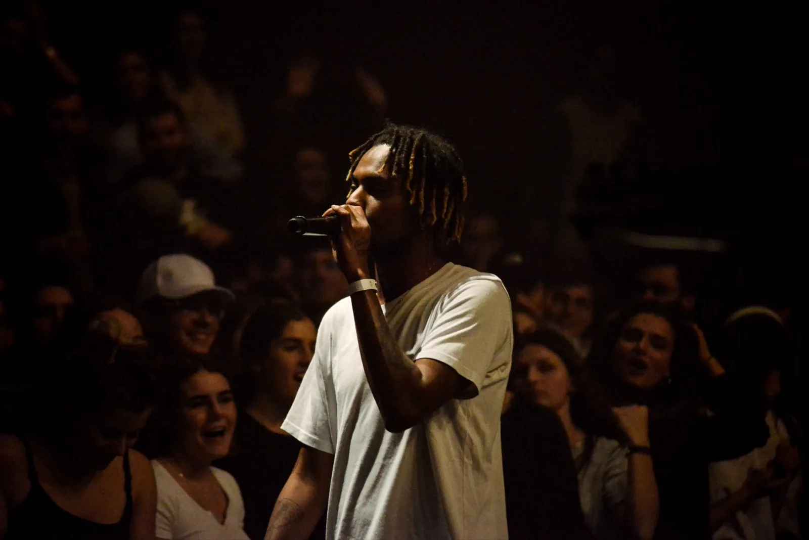 Rappeur debout au milieu de la foule, micro en main, regard tourné vers le haut, capté sous un éclairage de scène chaud.