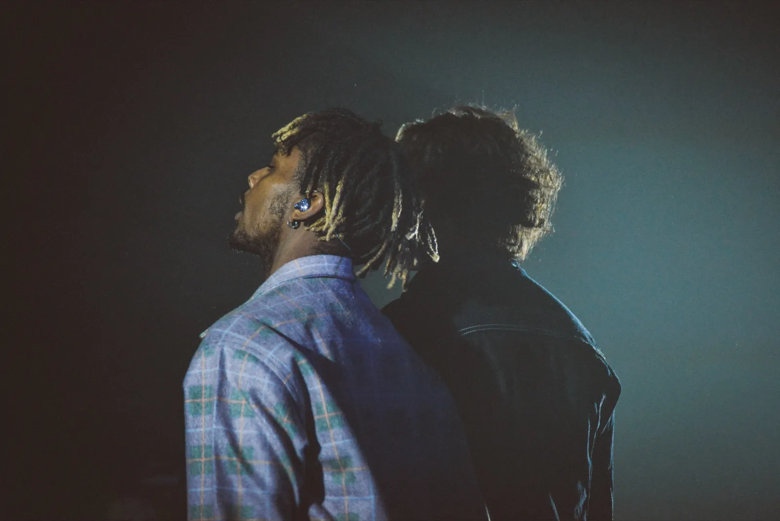 Deux rappeurs de dos, côte à côte sur scène, enveloppés d’une lumière bleutée dans une ambiance de concert intimiste.