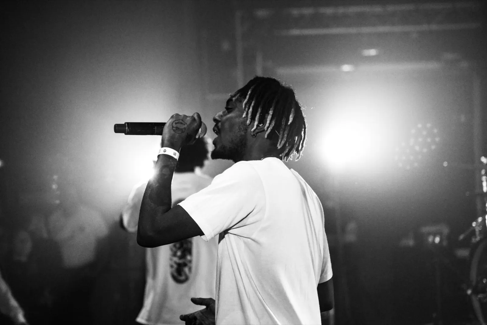 Vue de profil d’un rappeur sur scène, capté en pleine performance avec le micro tendu devant lui, projecteur intense en arrière-plan.