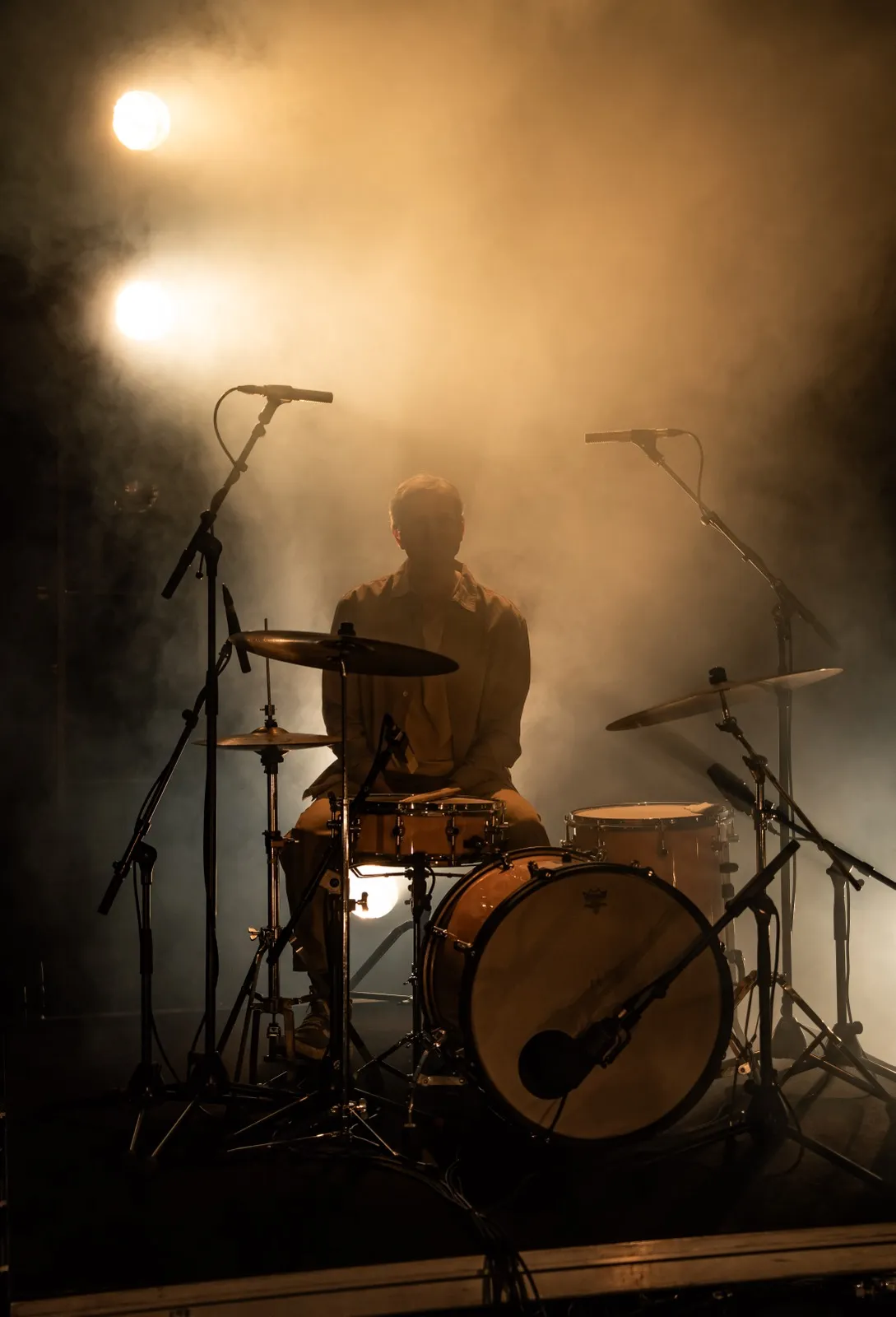 Batteur jouant dans un halo de lumière chaude, avec une scène embrumée et un jeu de clair-obscur marqué.