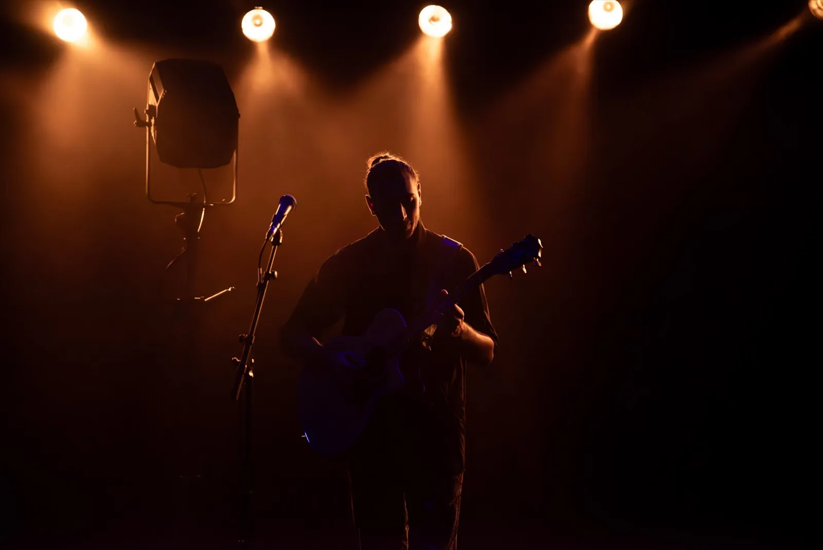 Silhouette d’un guitariste sous des projecteurs orangés, baigné dans une atmosphère de concert intimiste.