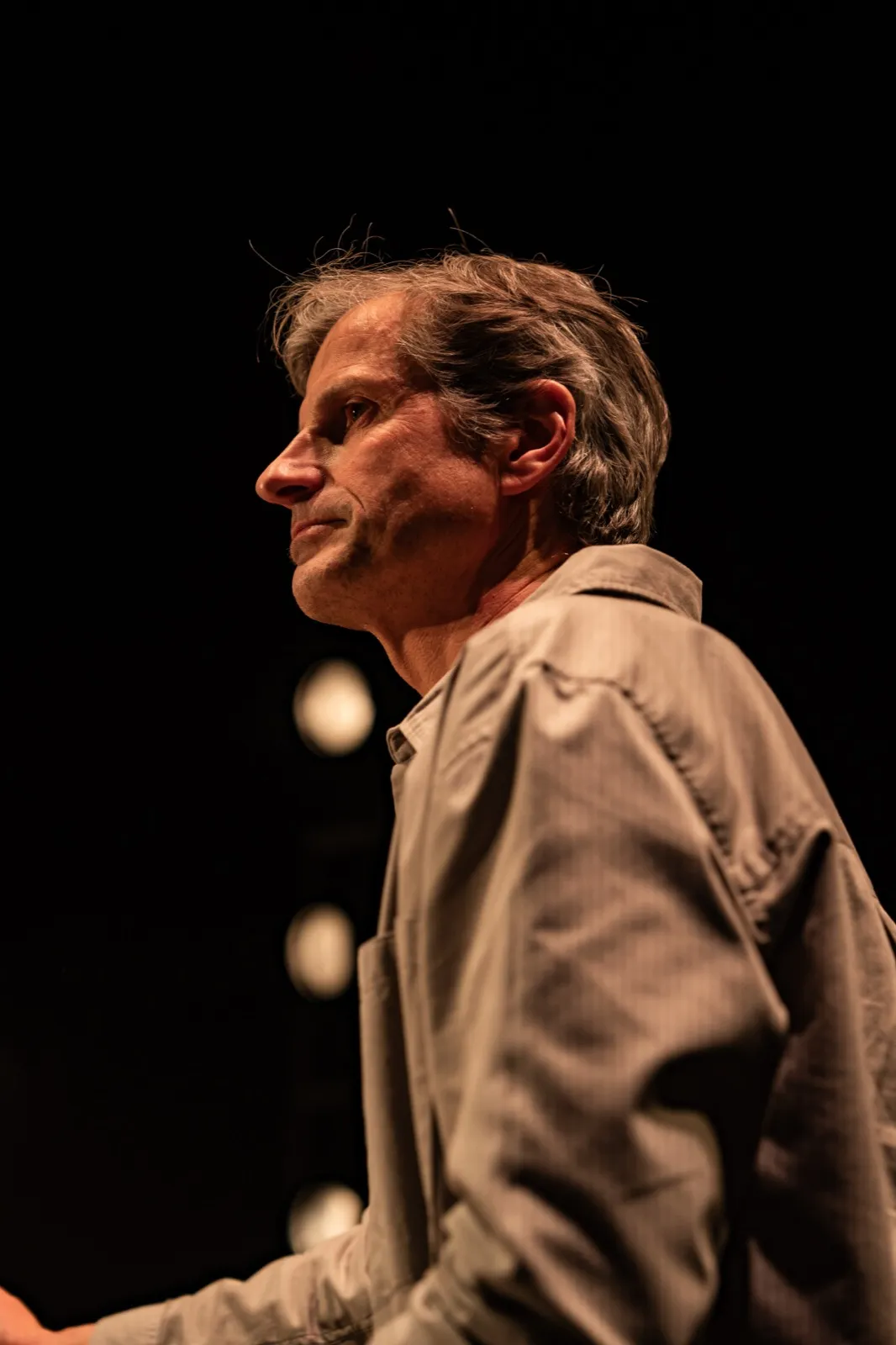 Portrait latéral d’un musicien plus âgé, capté sur scène dans une lumière chaude et tamisée.
