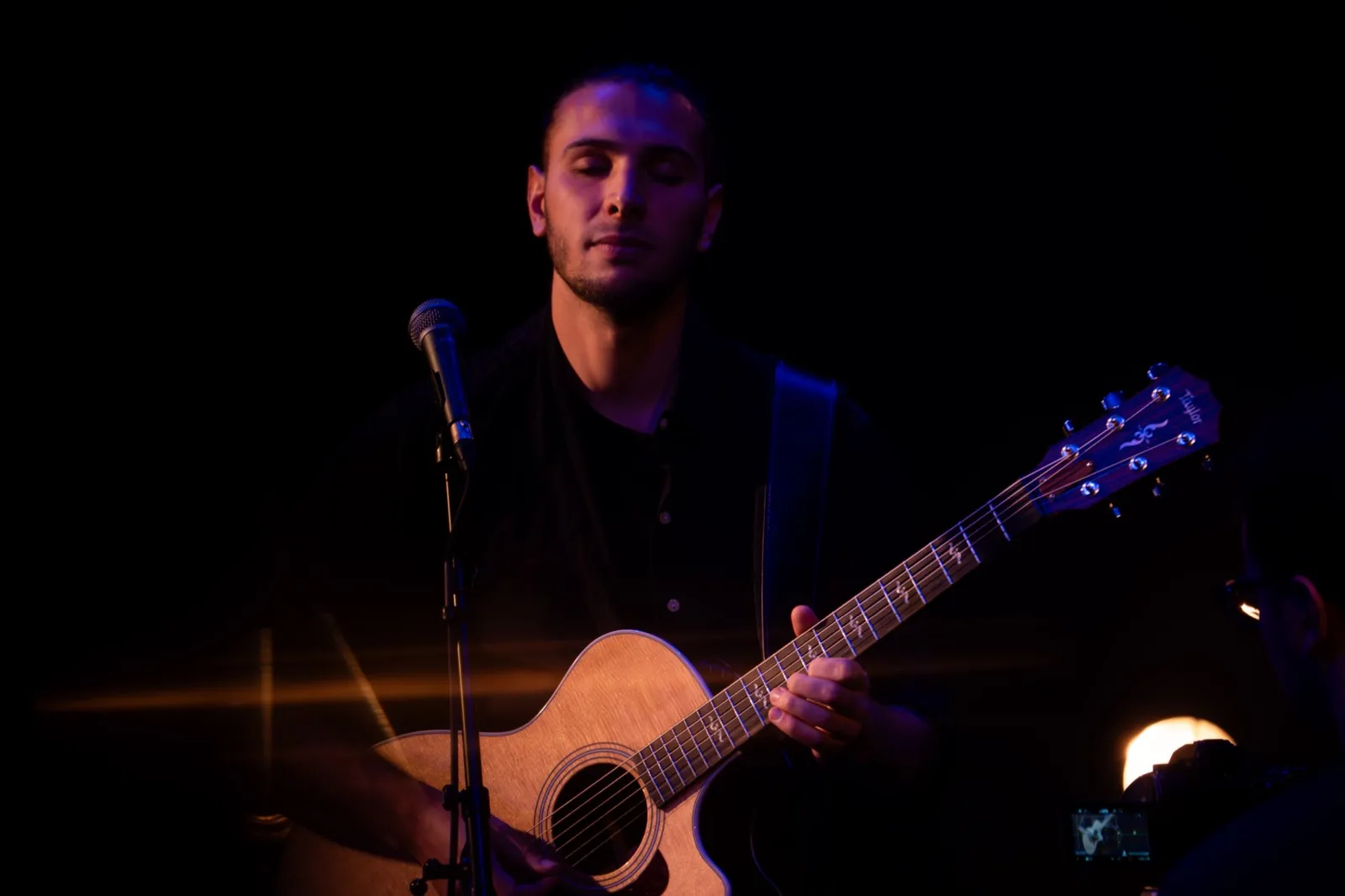 Artiste debout sur scène avec une guitare, éclairé faiblement dans une ambiance de concert intimiste.