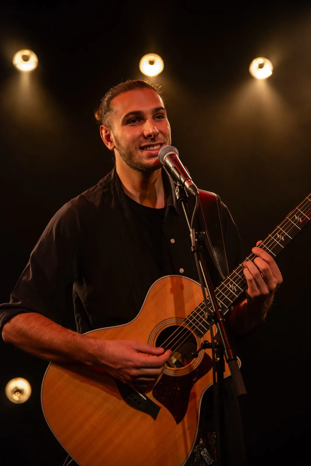 Musicien souriant sur scène avec une guitare acoustique, éclairé par des spots jaunes en arrière-plan.