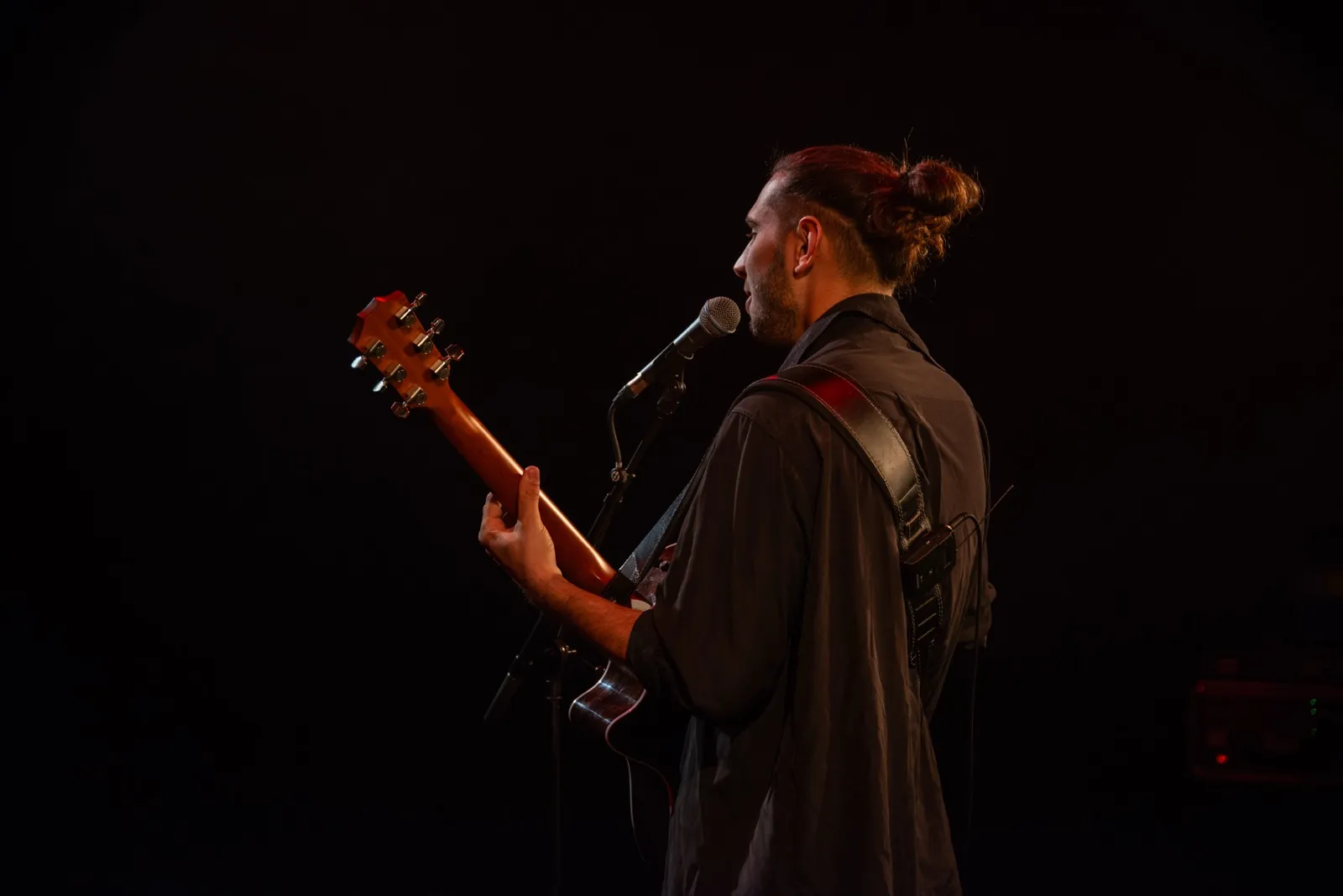 Chanteur vu de dos, jouant de la guitare sur scène dans une lumière tamisée, ambiance chaleureuse.
