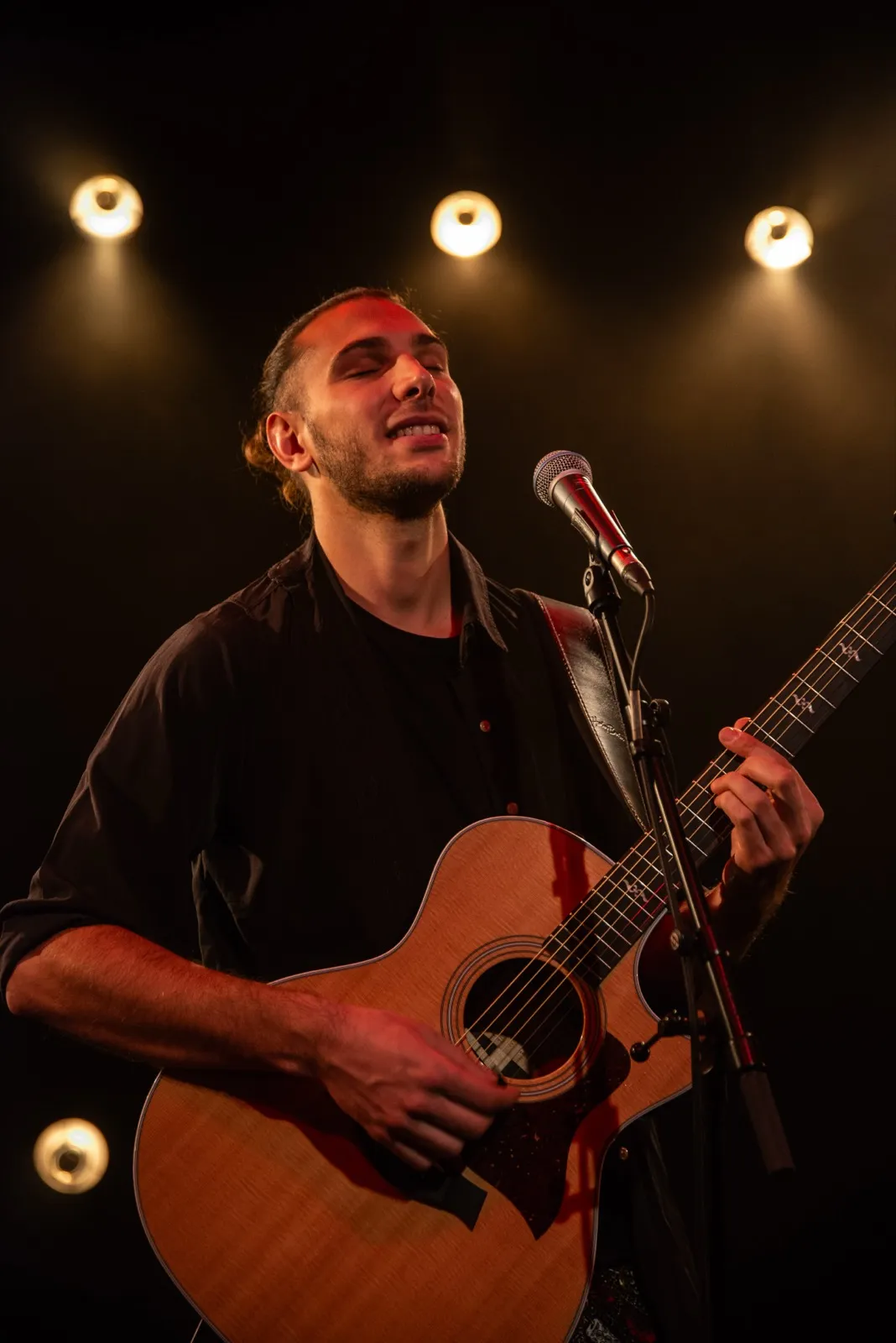 Guitariste souriant sur scène, tenant sa guitare acoustique, baigné dans une lumière orangée de concert.