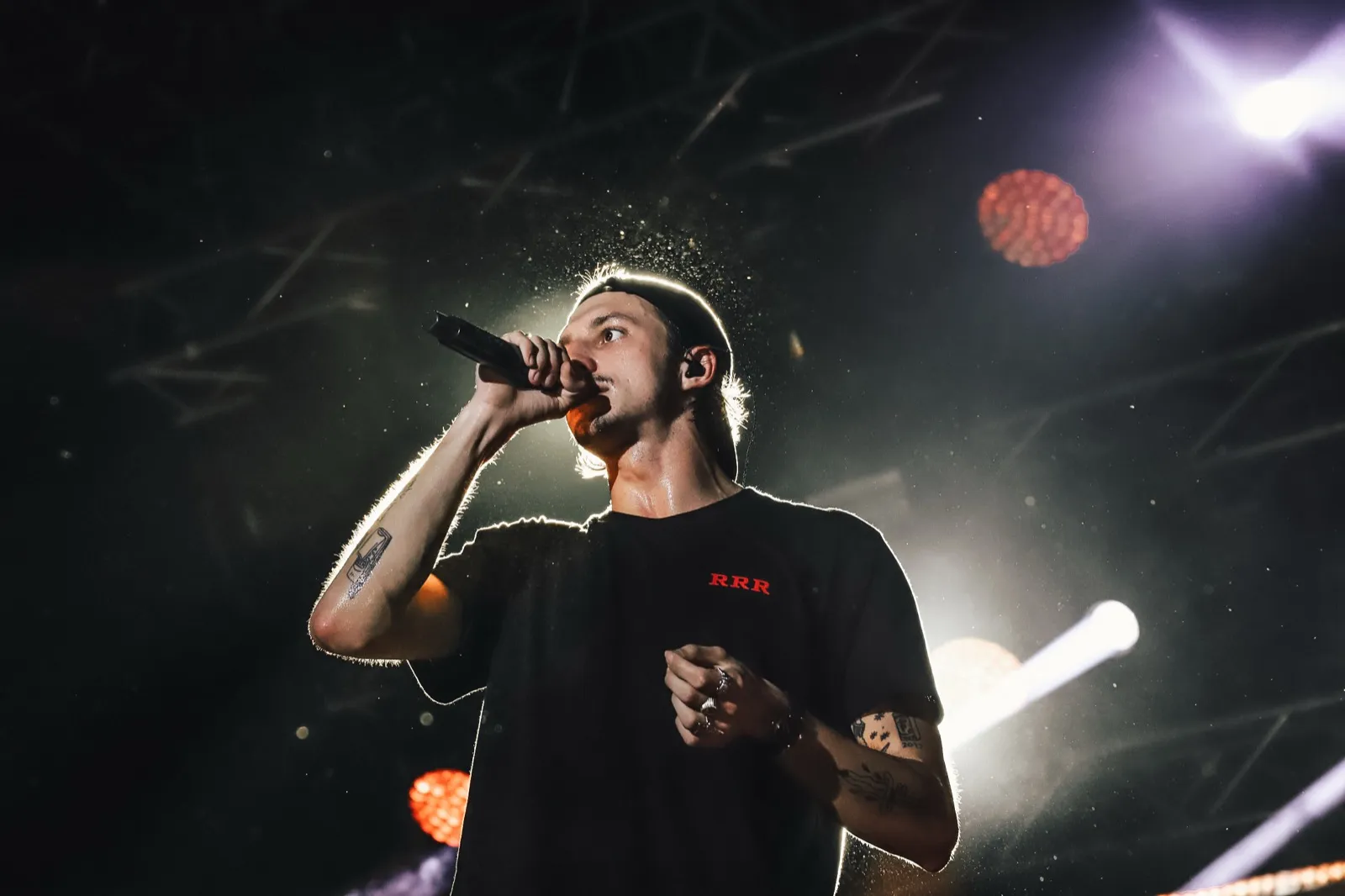 Un chanteur s’exprime sur scène, éclairé par des lumières violettes et blanches, regard dirigé vers le public.