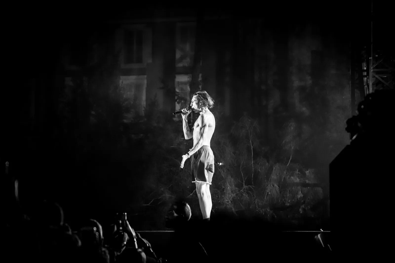 Un chanteur marchant sur scène, mis en lumière devant une façade d’immeuble, ambiance nocturne et intense.