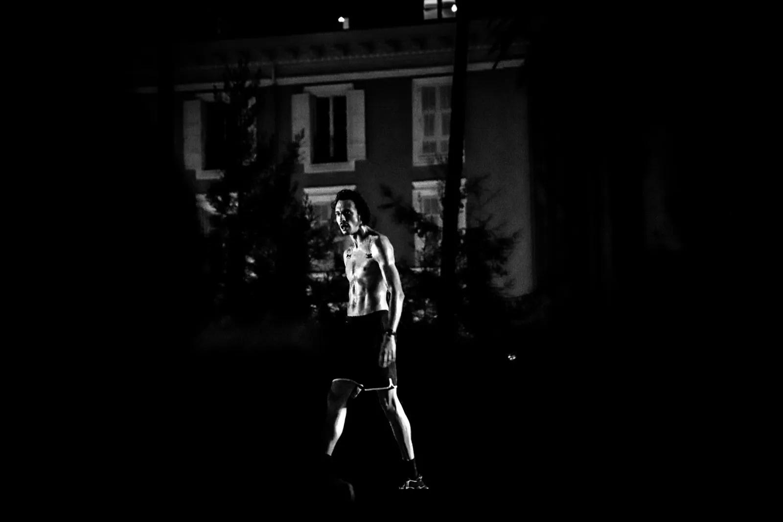 Un chanteur marchant sur scène, mis en lumière devant une façade d’immeuble, ambiance nocturne et intense.