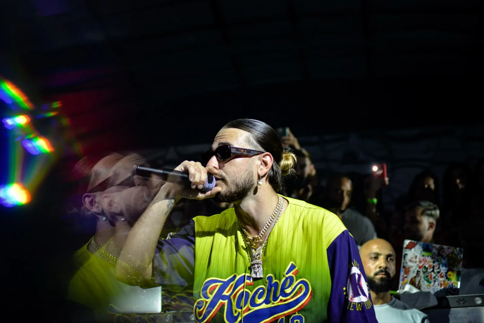 Un rappeur en pleine performance avec des lunettes de soleil et une chemise jaune vif, entouré par la foule dans une ambiance de concert int