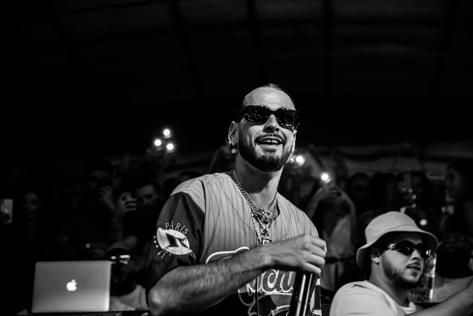 Un rappeur arborant un large sourire, habillé d’un maillot de baseball, capturé lors d’un concert intimiste en noir et blanc.