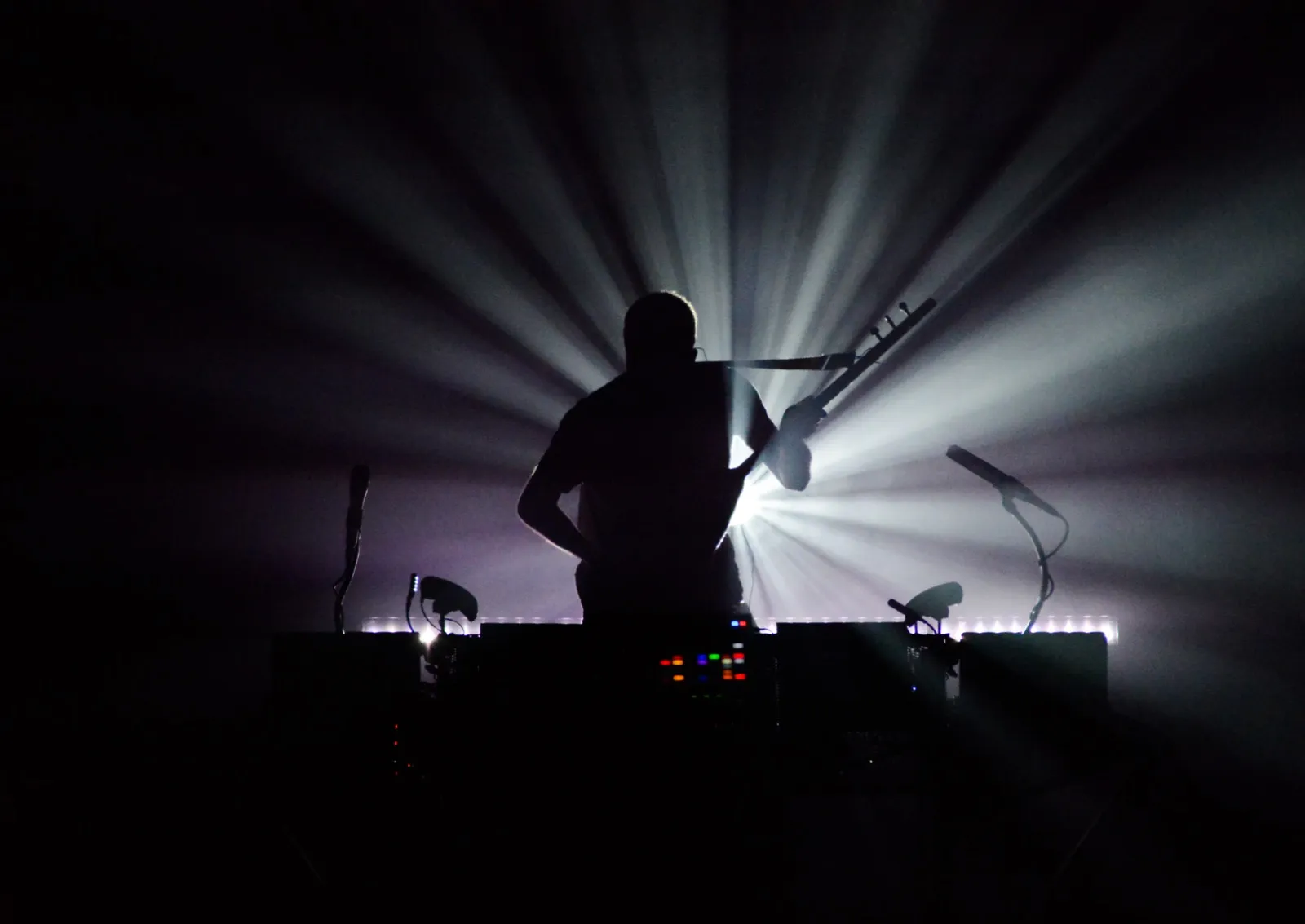 Silhouette d’un DJ sur scène, face à un public invisible, encadré de puissants faisceaux lumineux émanant de derrière lui.