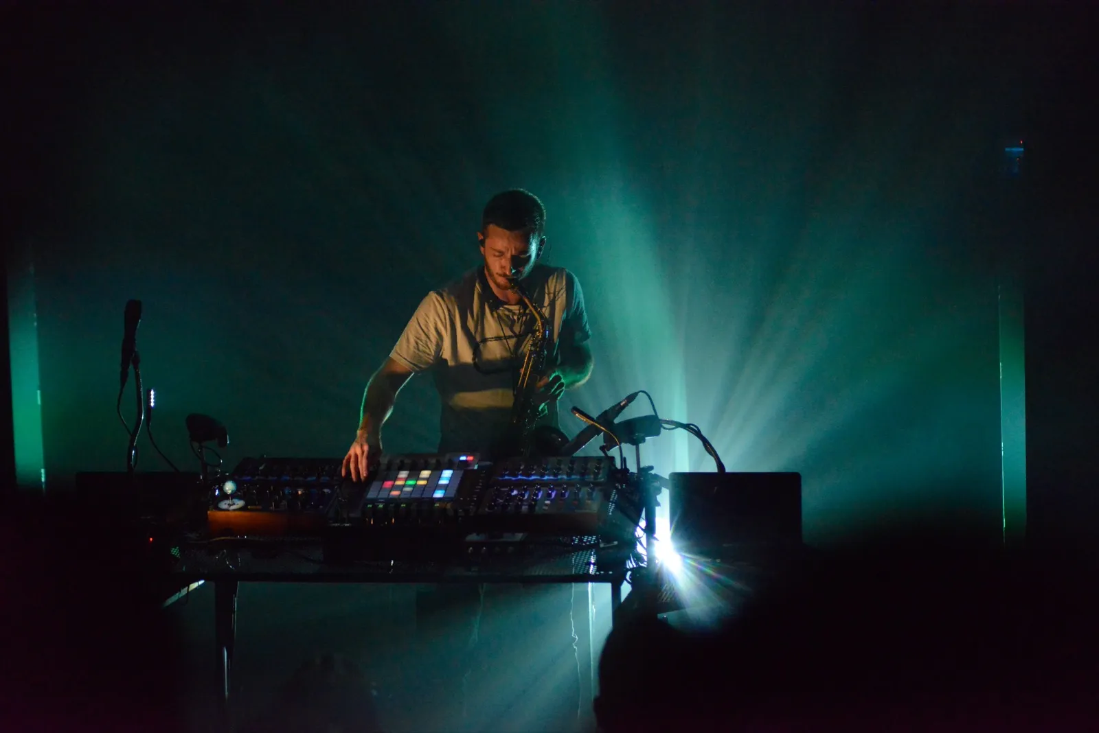Un DJ ou producteur électronique joue sur scène dans une lumière verte intense, entouré de matériel de production musicale.
