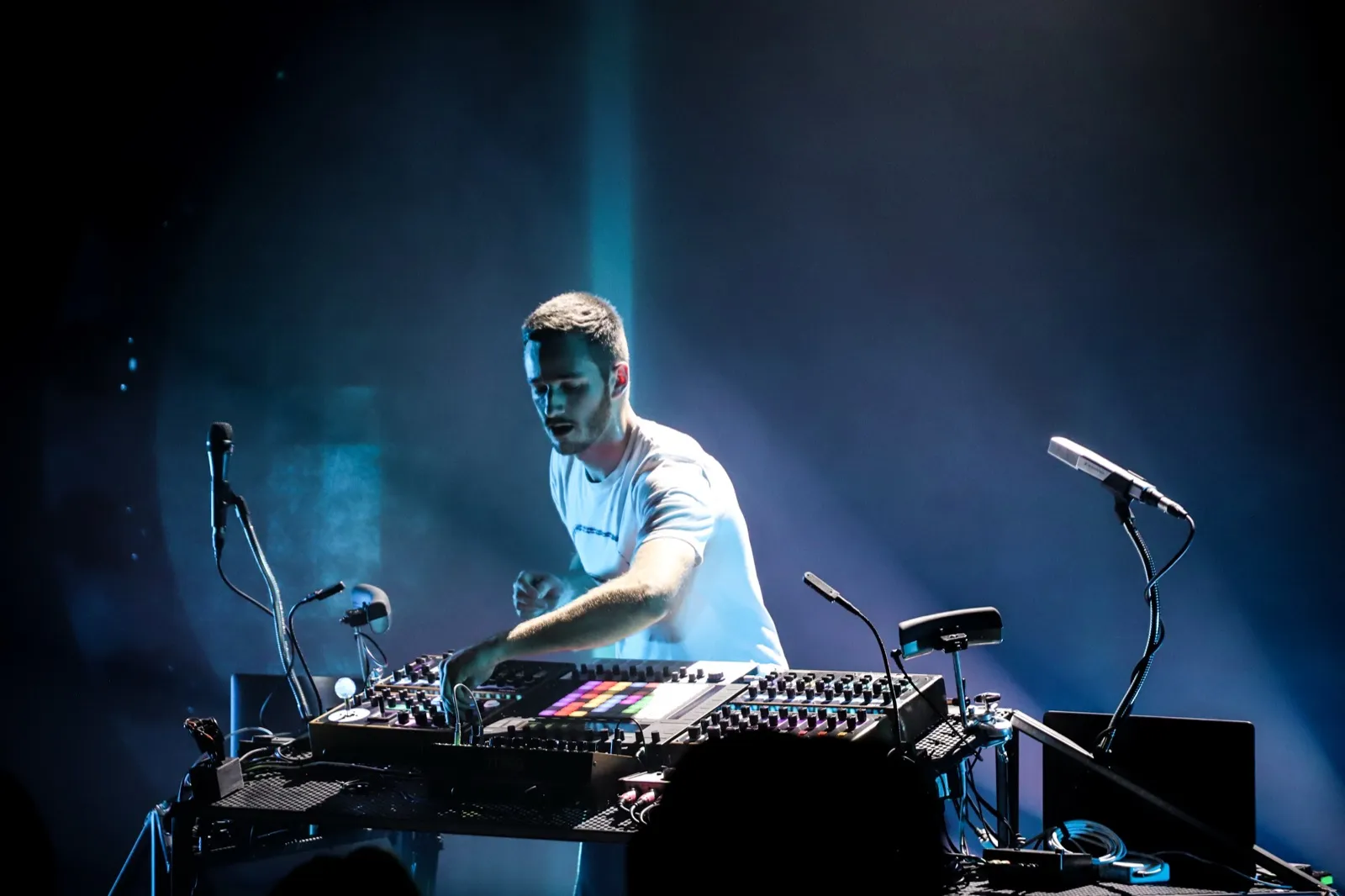 Un producteur en pleine performance live entouré de synthétiseurs et de câbles, baigné de lumière bleue et violette.