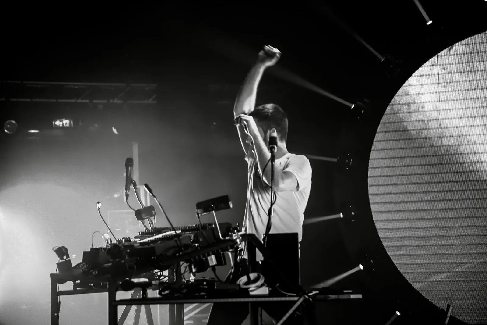 Un DJ en pleine performance, bras levé, derrière un setup électronique, éclairé par une lumière de scène dramatique.
