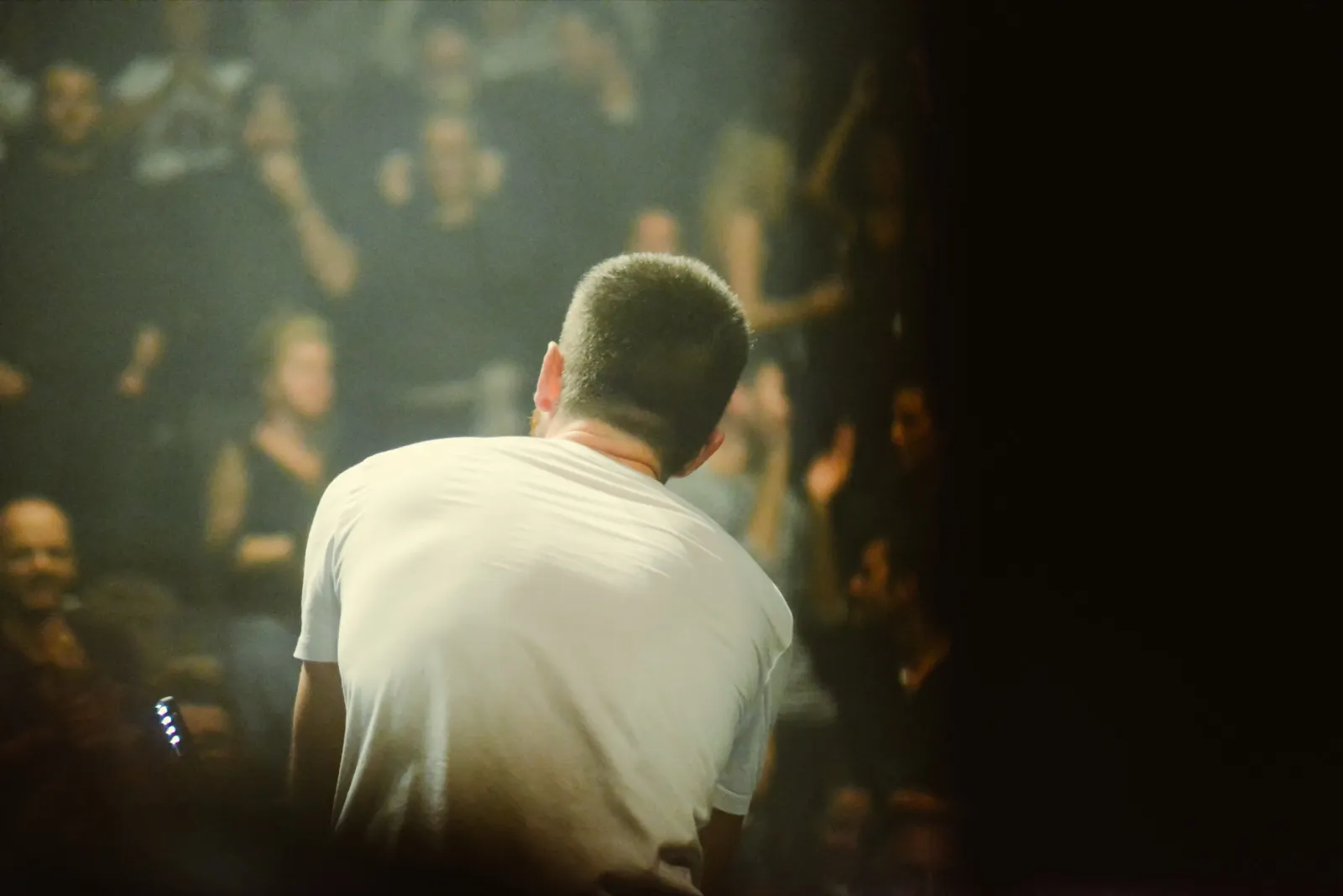 Un chanteur vu de dos, t-shirt blanc, s’adresse à une foule floue dans une lumière chaude et tamisée.