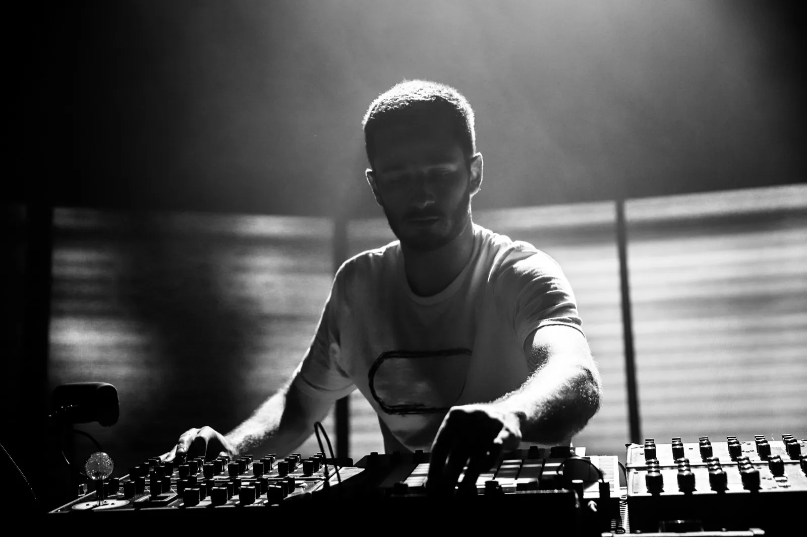 Un DJ en t-shirt clair joue sur scène, penché sur ses contrôleurs avec un éclairage zébré sur le fond.