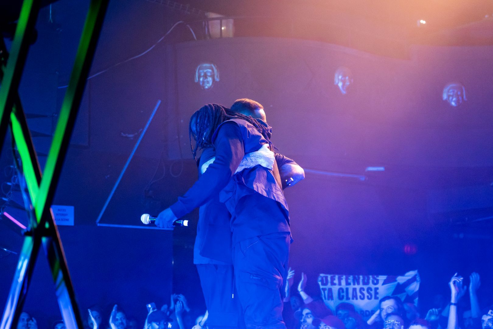 Moment intense de concert avec un rappeur serrant un proche sur scène, lumière bleu-violet et ambiance festive