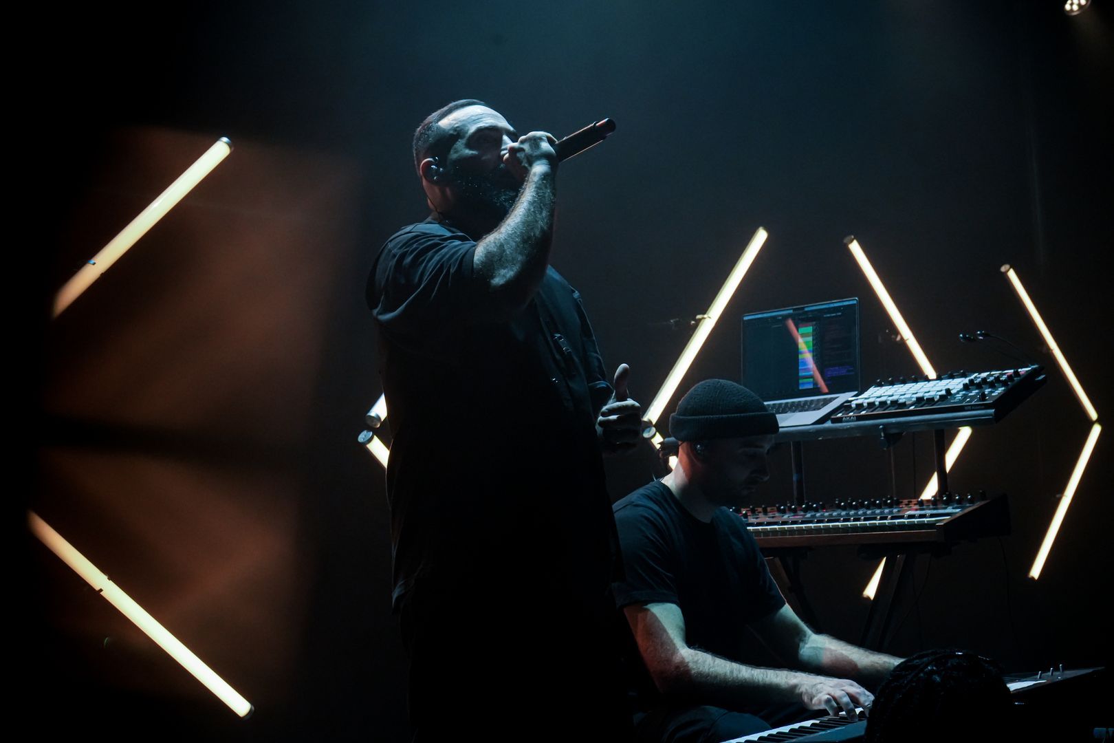 Chanteur de rap en live, micro en main, accompagné d’un claviériste, devant un fond lumineux stylisé