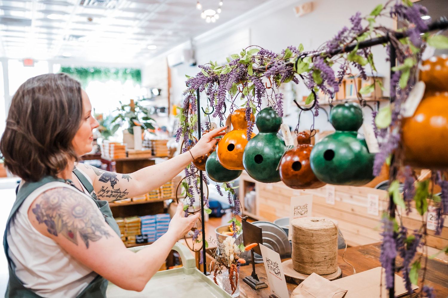 Person in white tank top arranging dried floral decor and green glass gourds on retail display shelf.