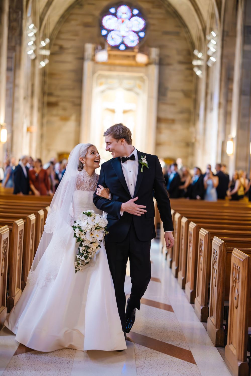 A bride and groom walk down the center aisle of a grand cathedral church with stained glass windows and wooden pews.