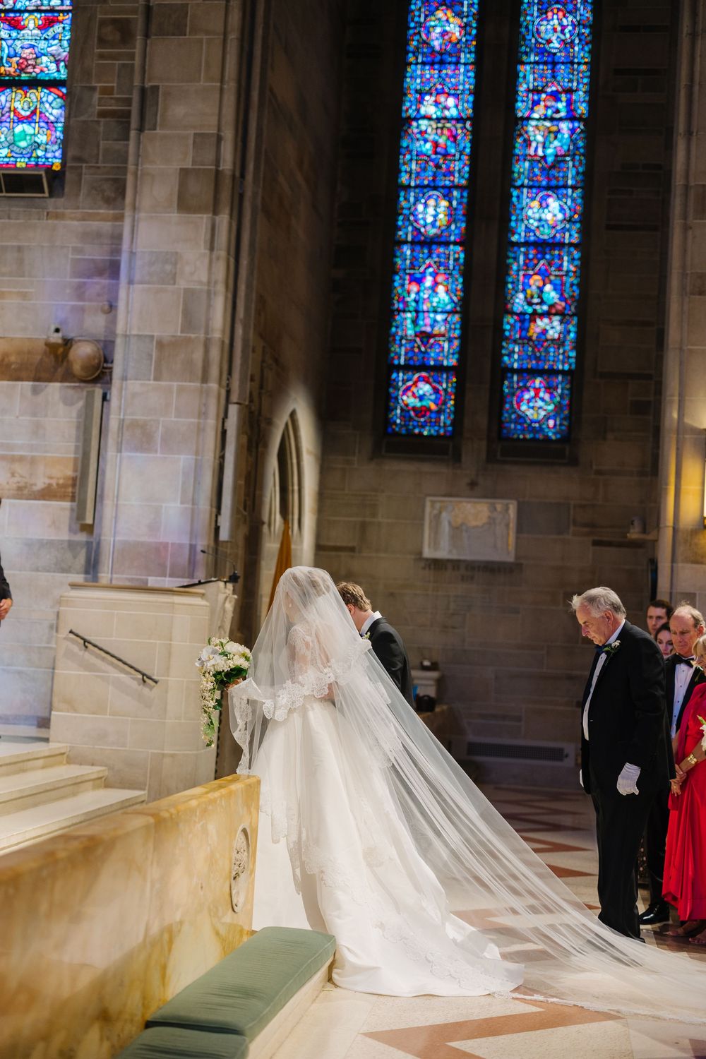 A bride in a flowing white dress with a long cathedral veil stands at church altar steps near stained glass windows.