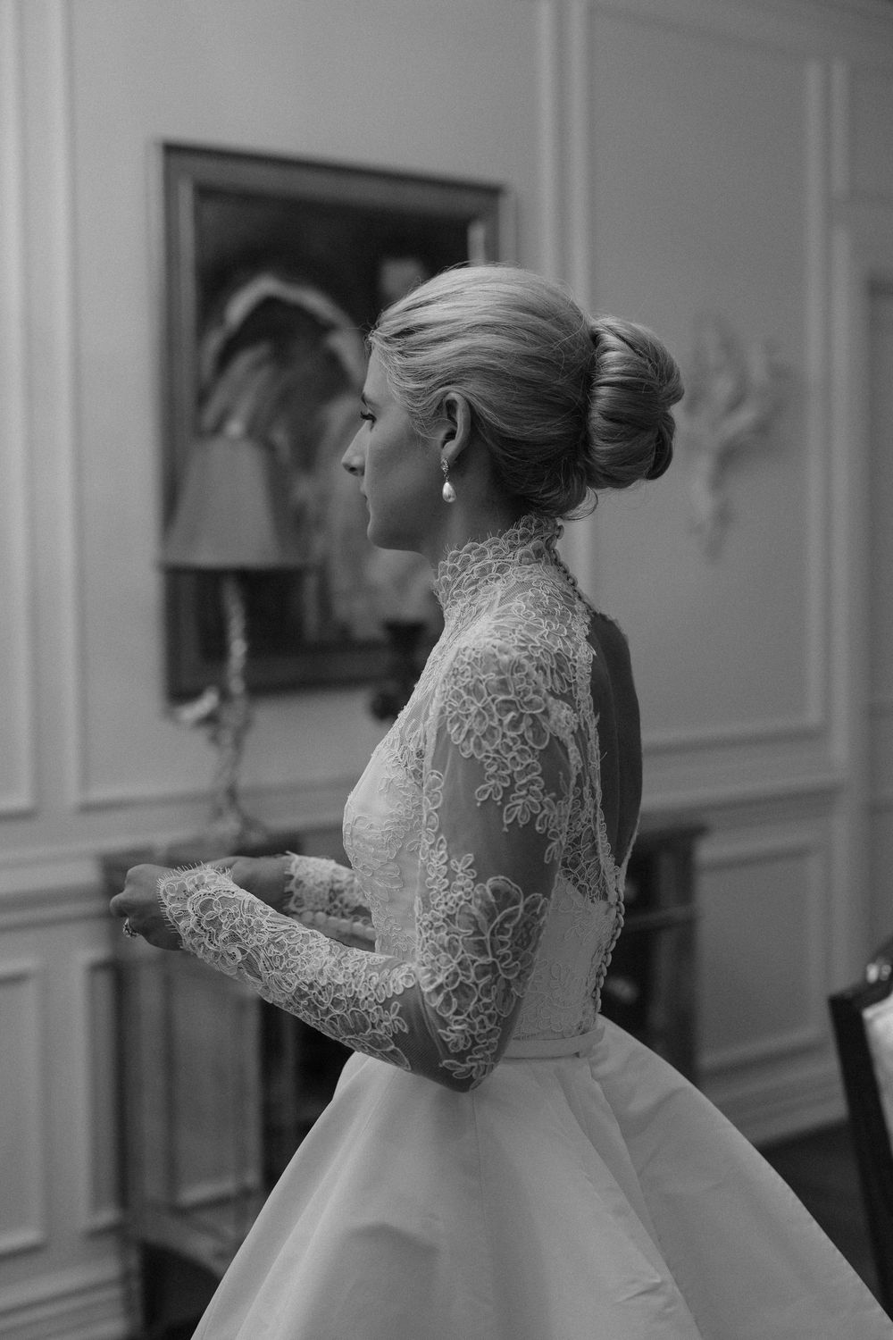 Black and white photo showing an elegant bridal gown with delicate lace sleeves and a high neckline, styled with a classic updo.