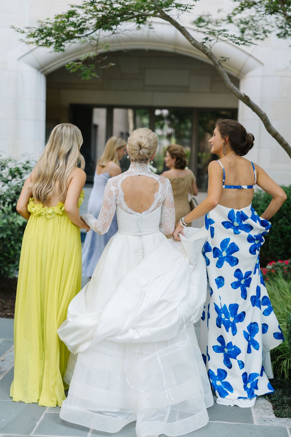 Bridesmaids in yellow and blue floral dresses stand with bride wearing classic white lace gown with keyhole back.