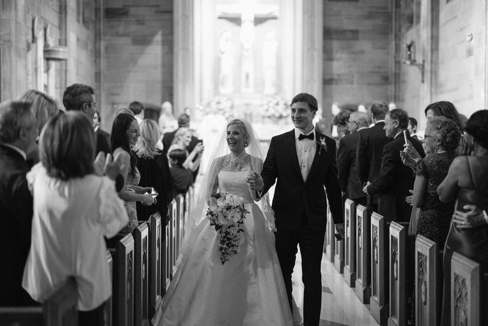 A wedding couple walks down the aisle as guests look on in a rustic church with light streaming through windows.
