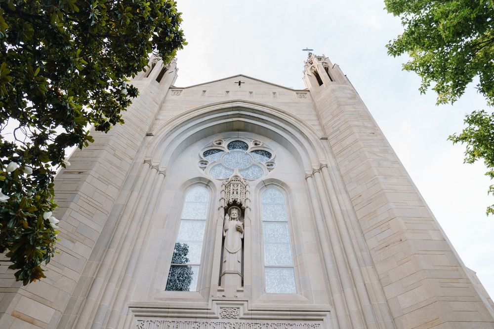 Looking up at a tall Gothic church facade with arched windows and religious statuary surrounded by green trees.