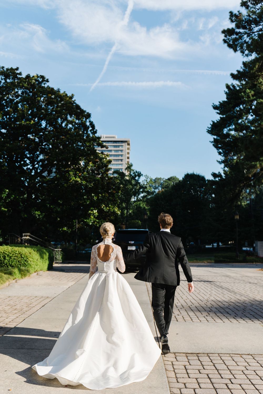 A couple in wedding attire walking on a paved pathway with tall trees and a high-rise building in the background.