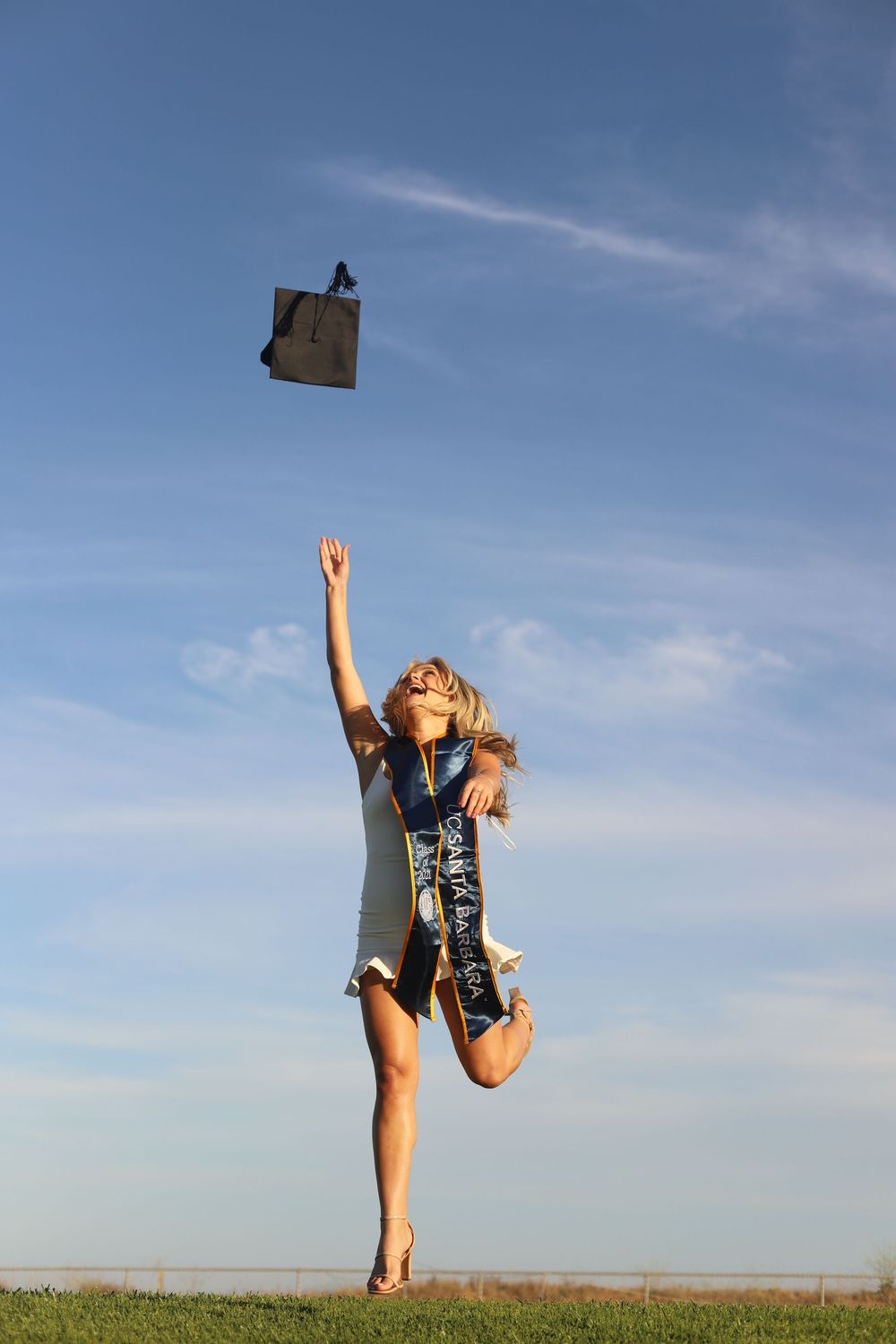 A graduate in a blue gown joyfully jumps and tosses their diploma case into the air against a sunny sky.