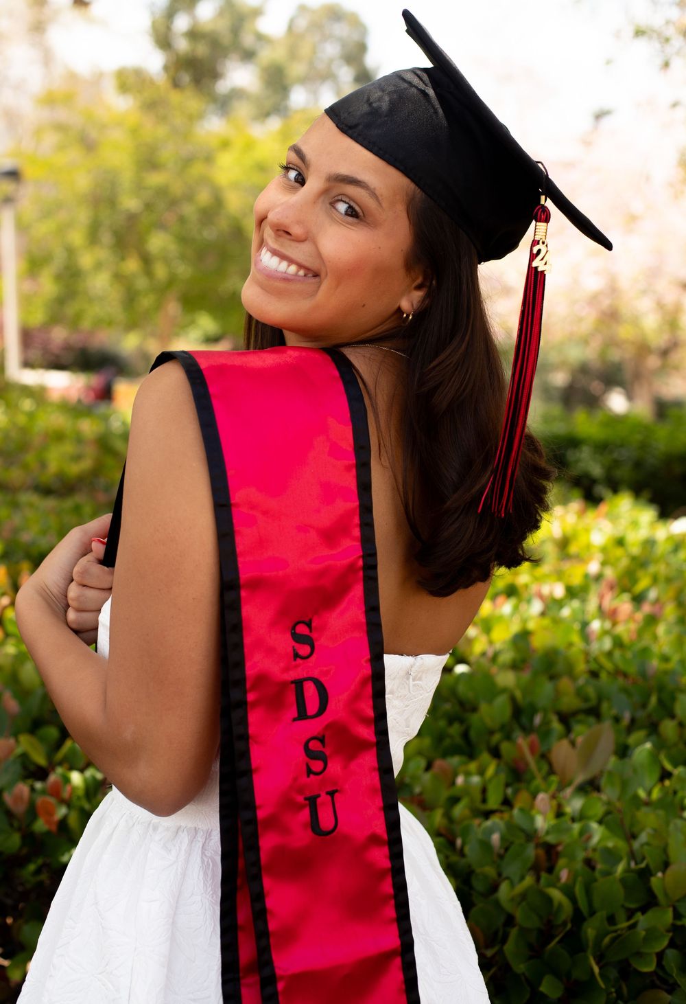 Graduate in cap and white dress wears a red SDSU sash while posing for a graduation photo outdoors.