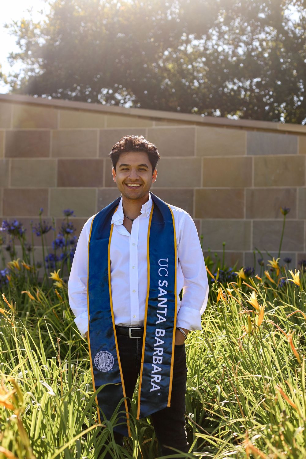 Graduate stands in a sunny garden wearing a white shirt and UC Santa Barbara graduation sash with purple flowers in background.