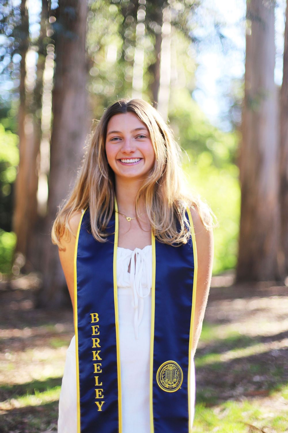 Graduate wearing a UC Berkeley graduation stole and white top smiles in a sunny forest setting.