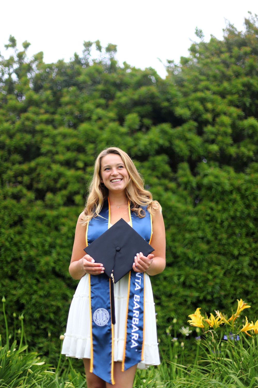 Graduate in white dress holds mortarboard and wears blue graduation stole while standing outdoors with green foliage background.